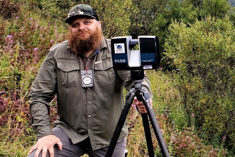 A man stands in a forested area holding a LiDAR camera on a camera stand.