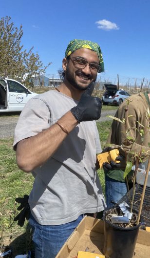  a man holds a thumb up standing near a potted plant