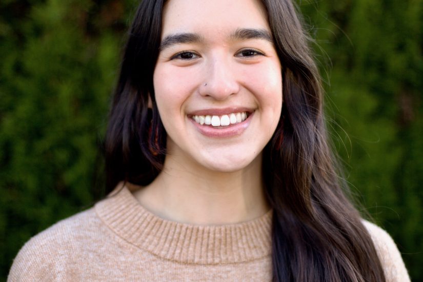 A woman with long dark hair and a beige sweater in front of foliage.