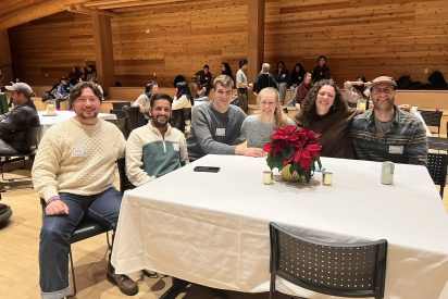 Students sit at a table at a holiday party with a wood paneled background and a poinsettia in the foreground.