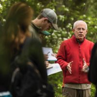 Man in a red jacket instructs students outdoors with trees in the background.