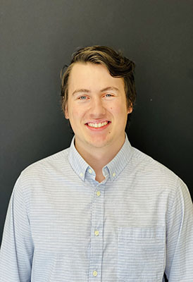 A man in a blue collared shirt stands in front of a black background.