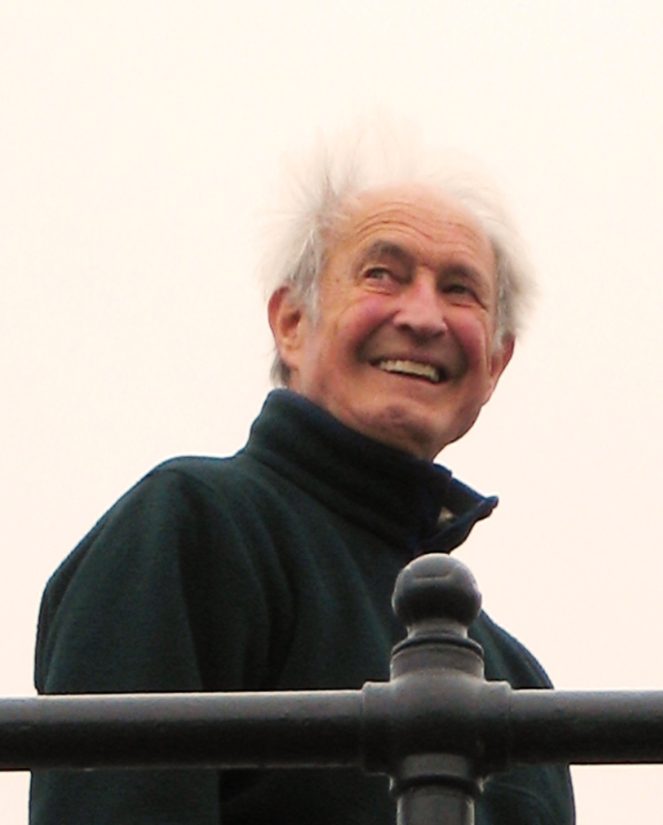 An old man with gray hair wearing a black fleece jacket smiles.
