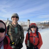 Three people dressed in winter clothes stand in the snow with mountains and trees in the background.