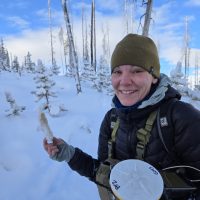 A woman dressed in snow clothes holds up a rabbits foot with trees and a blue sky in the background.