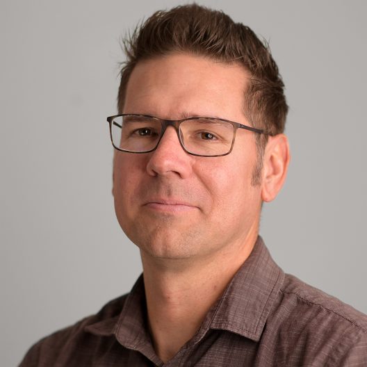 A man in tortoise shell glasses and a brown shirt poses for a headshot.