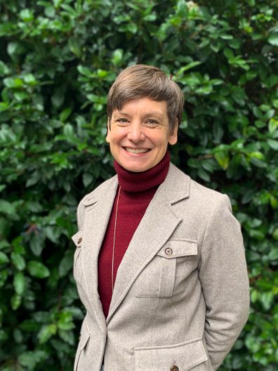 A woman in a burgundy turtleneck and a cream blazer smiles for a headshot. Leafy trees are in the background.