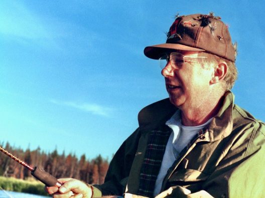 A man in a fishing hat covered in lures casts a fishing pole against a bright blue sky. 